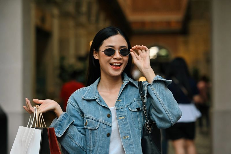 A fashionable woman in a denim jacket smiles while adjusting her sunglasses, holding shopping bags outdoors, a shopping spree in the city.