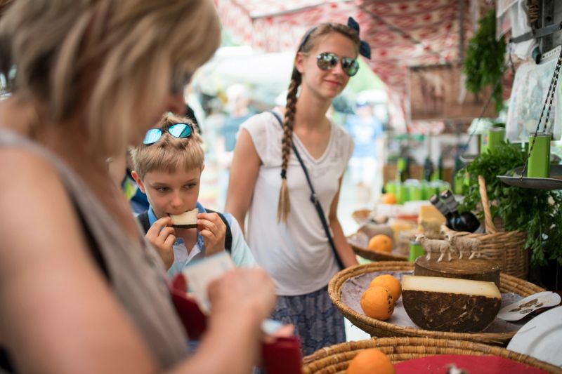 A multigenerational family pauses at a bustling market stall where a young boy takes of bite of a piece of cheese, while his sister in sunglasses watches with curiosity. Their grandmother stands close by, guiding them through the display of large cheese wheels, fresh oranges, herbs, and bottles of local products. Shot with Nikon D850