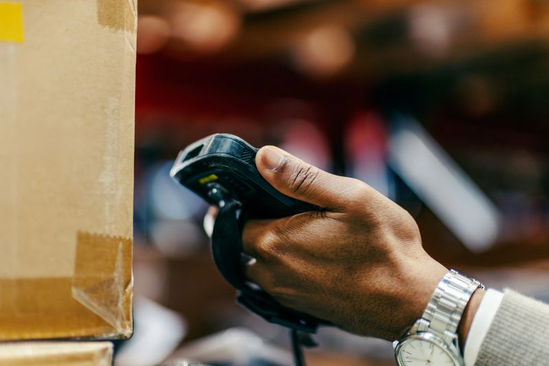 Worker hand holding a barcode scanner, efficiently scanning a brown cardboard box in a bustling logistics warehouse, ensuring accurate inventory management and quick delivery preparation