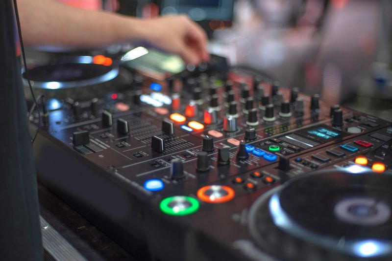 A high-angle close-up of a DJ's hand adjusting a professional mixing console during a live set. Featuring glowing buttons and a soft bokeh background with ample copy space, capturing the energy of nightlife and electronic music production