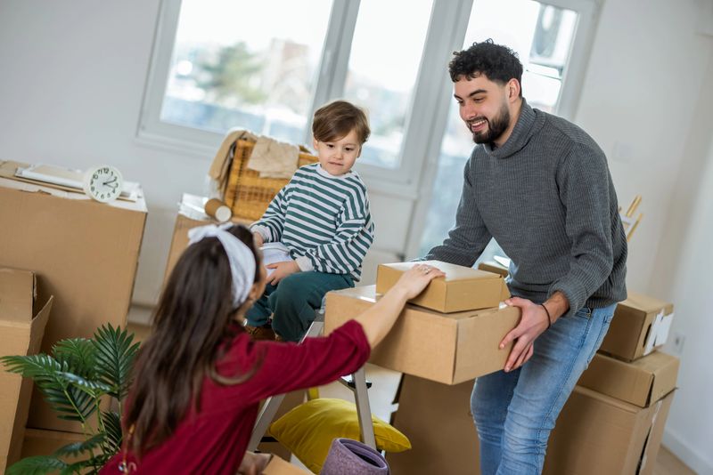 A young mother, father, and their small son are all carrying moving boxes across their bright, empty living room. The parents are lifting larger boxes while the toddler proudly carries a small one, mimicking his parents.