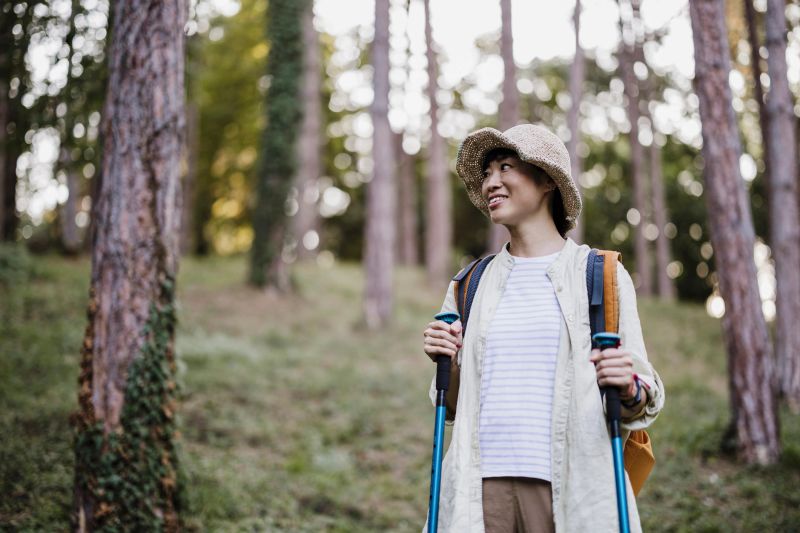 Portrait of a Japanese woman hiking outdoors, wearing a hat and backpack and holding trekking poles. Active female traveler enjoying nature, adventure, and a healthy lifestyle. Concept of hiking, outdoor recreation, eco tourism, and travel in nature.