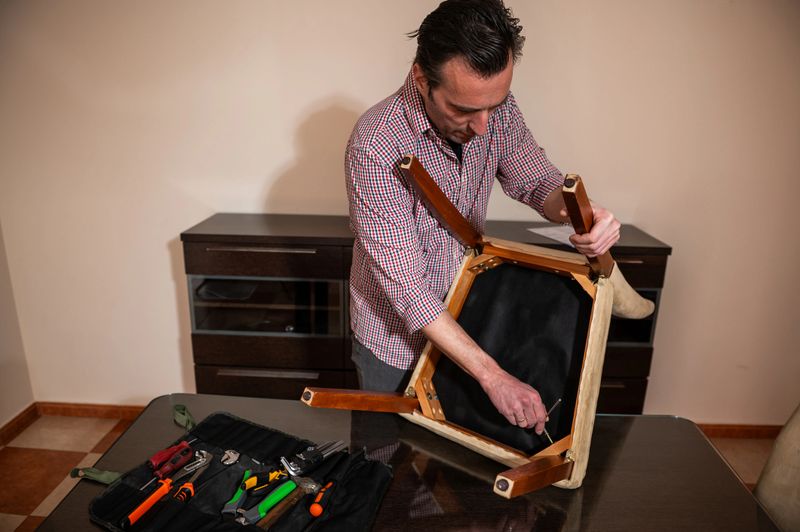 Middle-aged man repairs a wooden chair upside down on a dining table at home, using hand tools from a toolkit, illustrating DIY repair, home maintenance and hands-on craftsmanship.