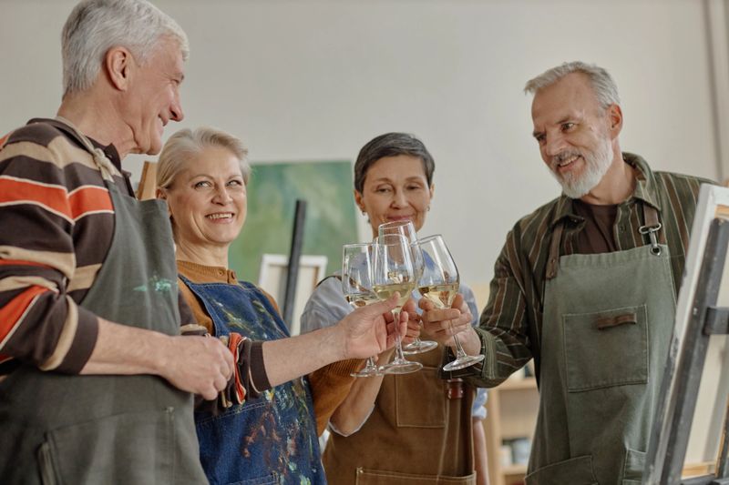 Group of senior Caucasian men and women standing together holding wine glasses and smiling during art class, wearing aprons and engaging in creative educational activity