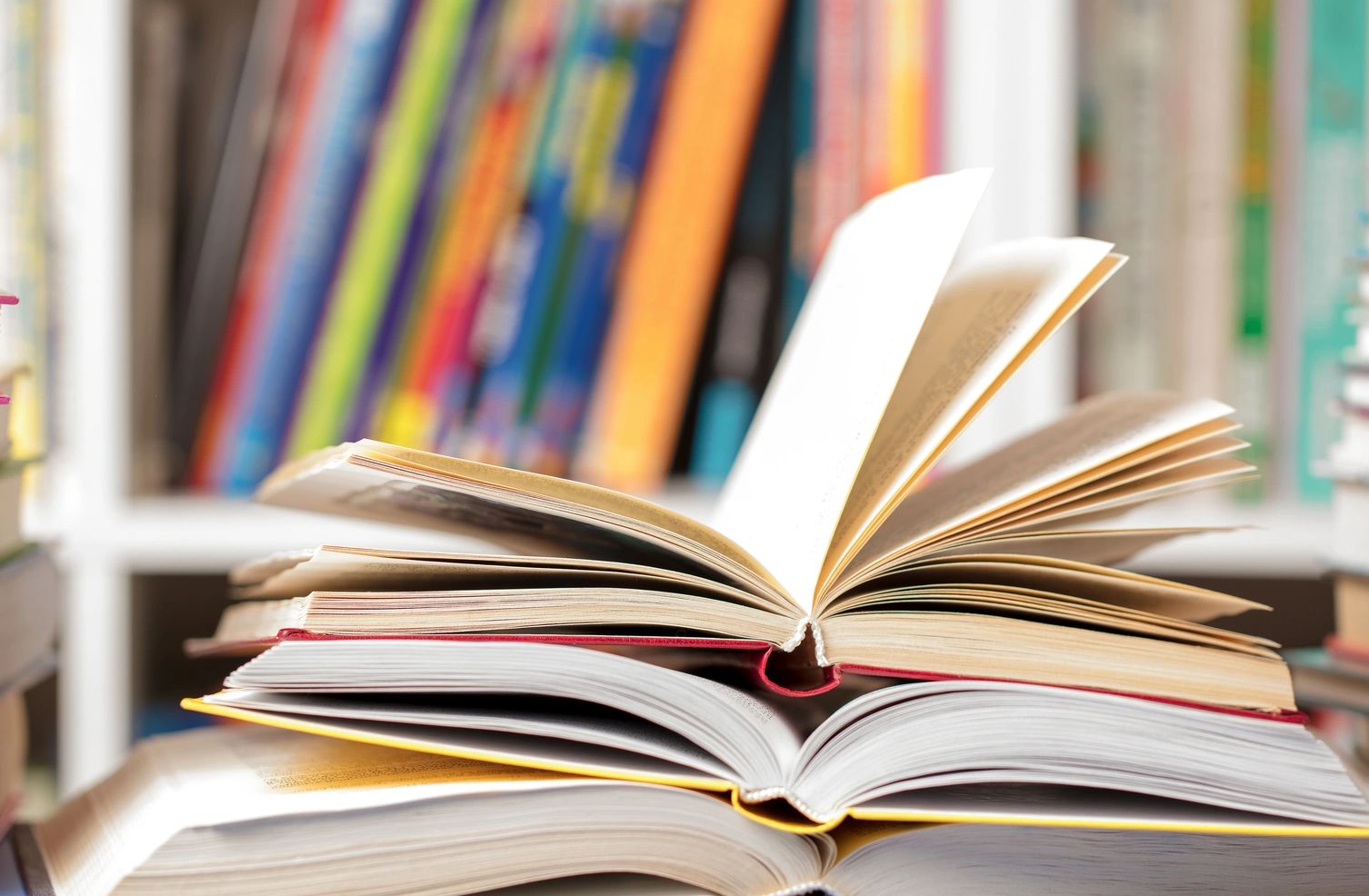 Stack of open books with colorful bookshelf in background.