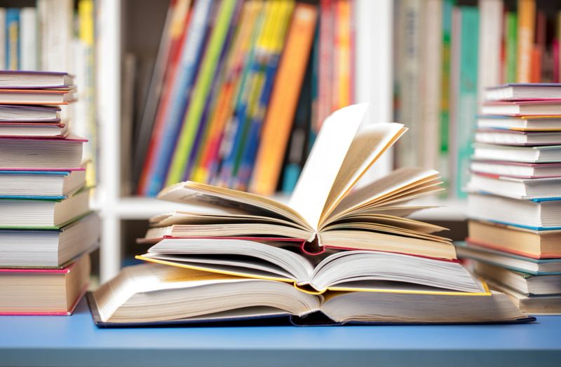 A DSLR photo of books at the library. Several books are arranged on a table with some open and others closed. The books have different colored spines. Blurred shelves of more books are in the background. Space for copy.