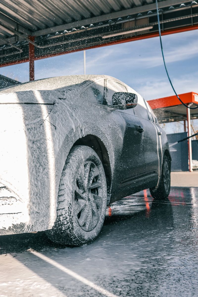 A black car covered in white foam at a car wash, with water spraying on it, creating a clean and refreshing look.