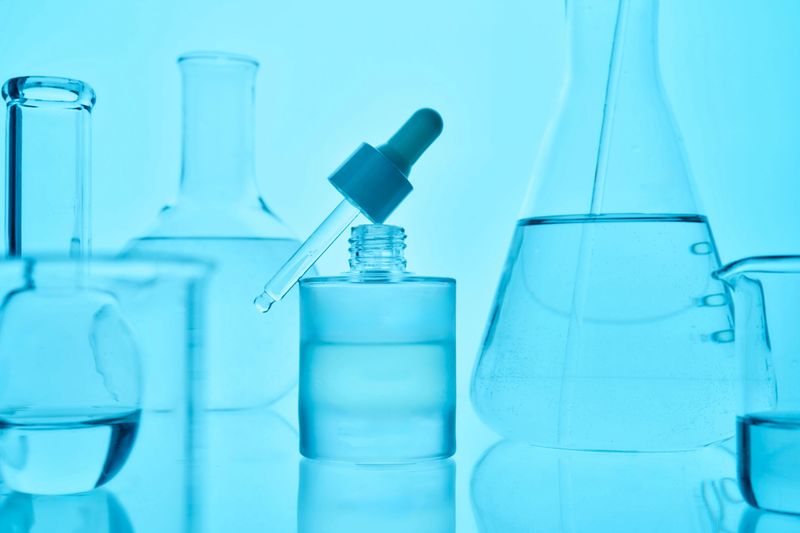 Skincare product dropper bottle with surrounding laboratory glassware containing clear liquid, showing scientific cosmetic development