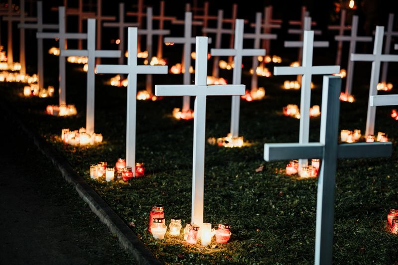 Rows of white crosses illuminated by red and white candles, creating a serene and solemn atmosphere at a nighttime memorial. The scene reflects respect, remembrance, and unity in tribute
