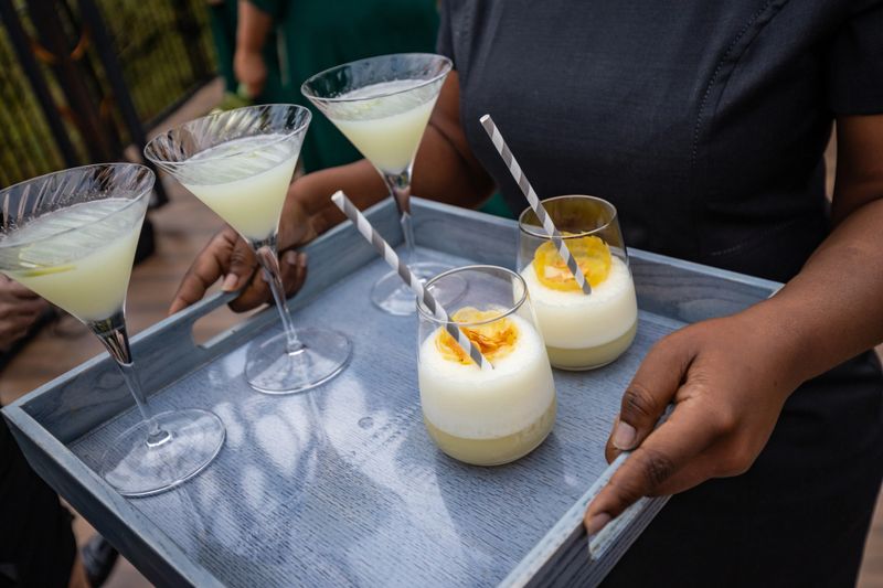 An elegant display of creamy cocktails and citrus drinks served on a tray by a server, featuring martini and glassware with striped straws and garnishes, ideal for a festive outdoor gathering