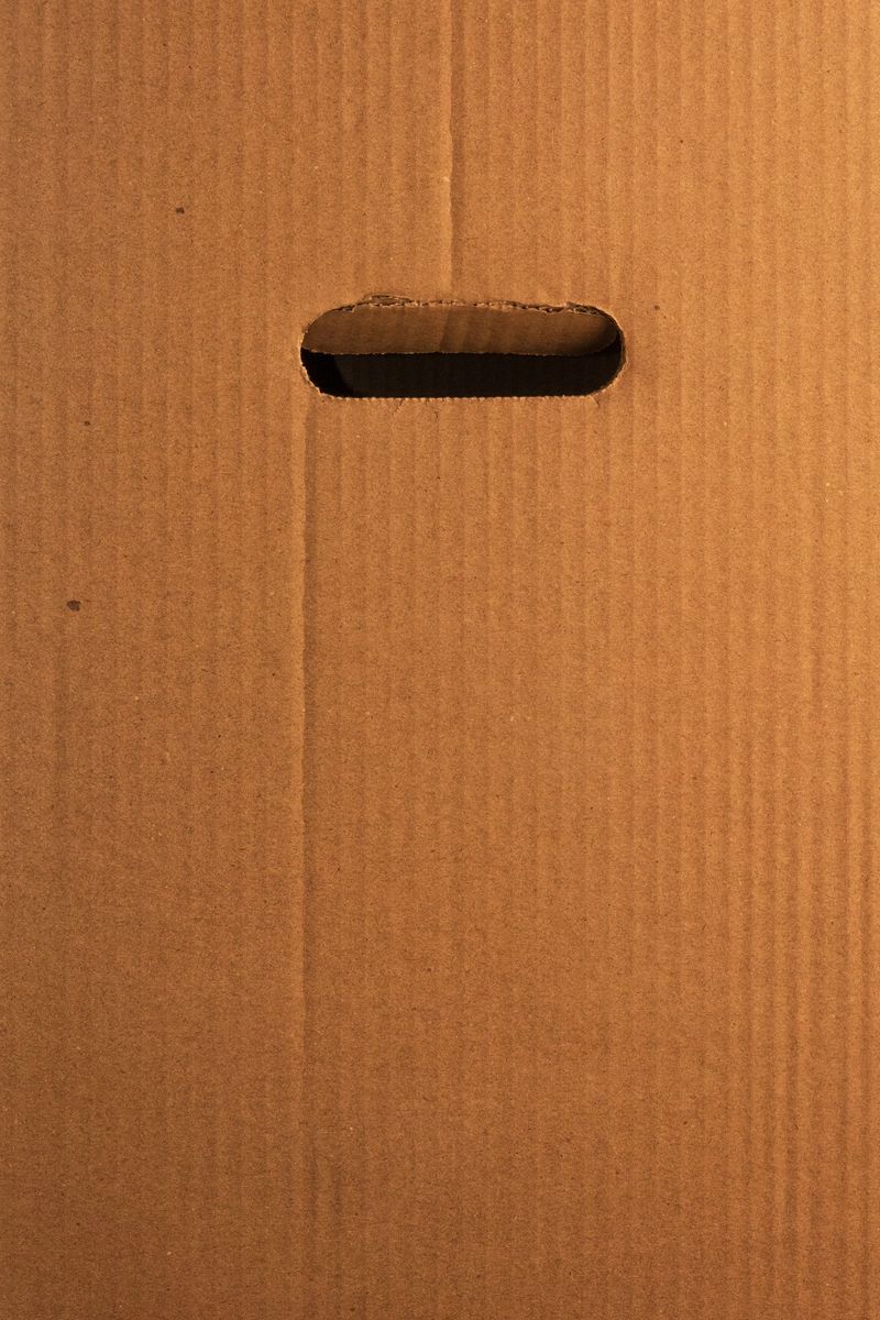 Close-up of a brown corrugated cardboard box with a handle. The minimalist, textured surface shows vertical lines. Warm, neutral colors. Ideal for shipping, moving, e-commerce, recycling concepts.