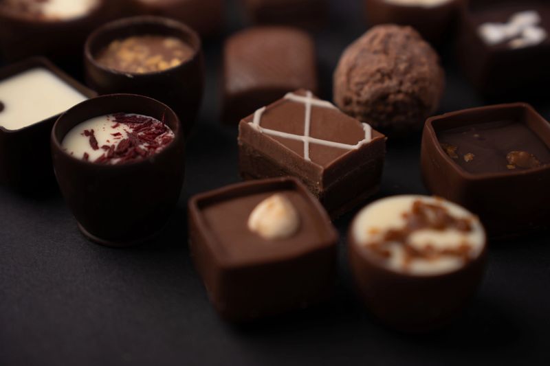 close-up of assorted gourmet chocolates and truffles on a matte black surface, highlighting rich brown and cream tones, glossy ganache, nut shavings and caramel, with a moody elegant feel, depth