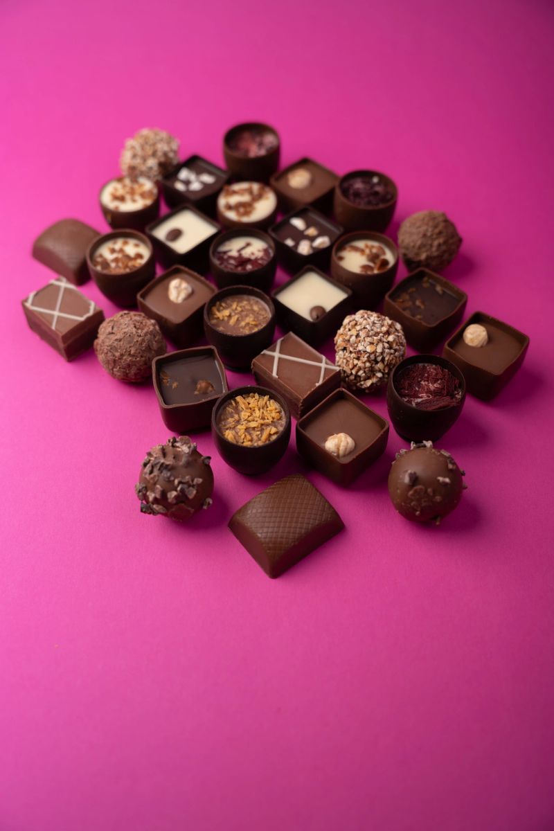 assorted gourmet chocolate pralines and truffles arranged in a scattered grid on a vivid magenta background, close-up shallow-depth composition showcasing varied textures, nuts and creamy fillings