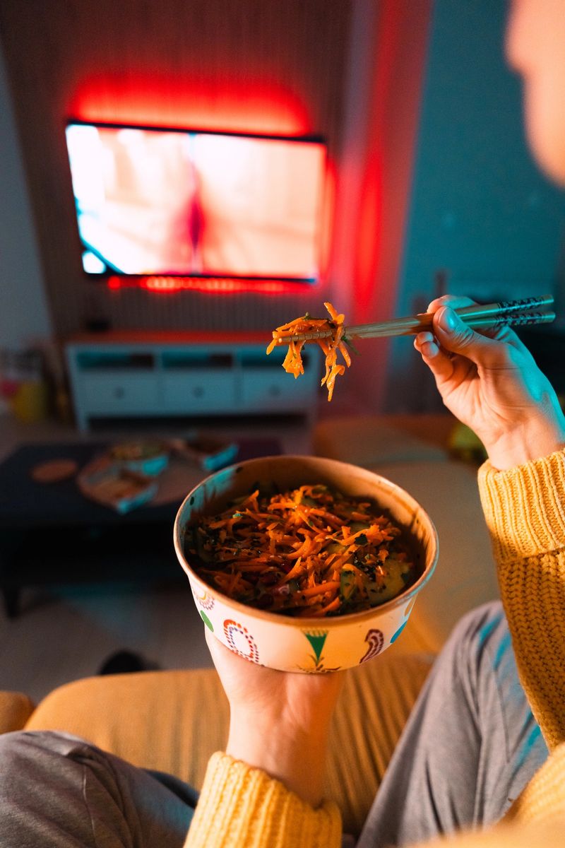 A woman lifts a piece of Asian salad with chopsticks while watching TV at home, showing healthy takeaway, fresh ingredients, and casual indoor dining.