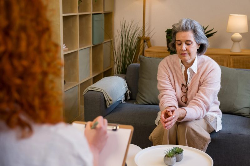 Senior woman sitting on couch with eyes closed, receiving support from a therapist