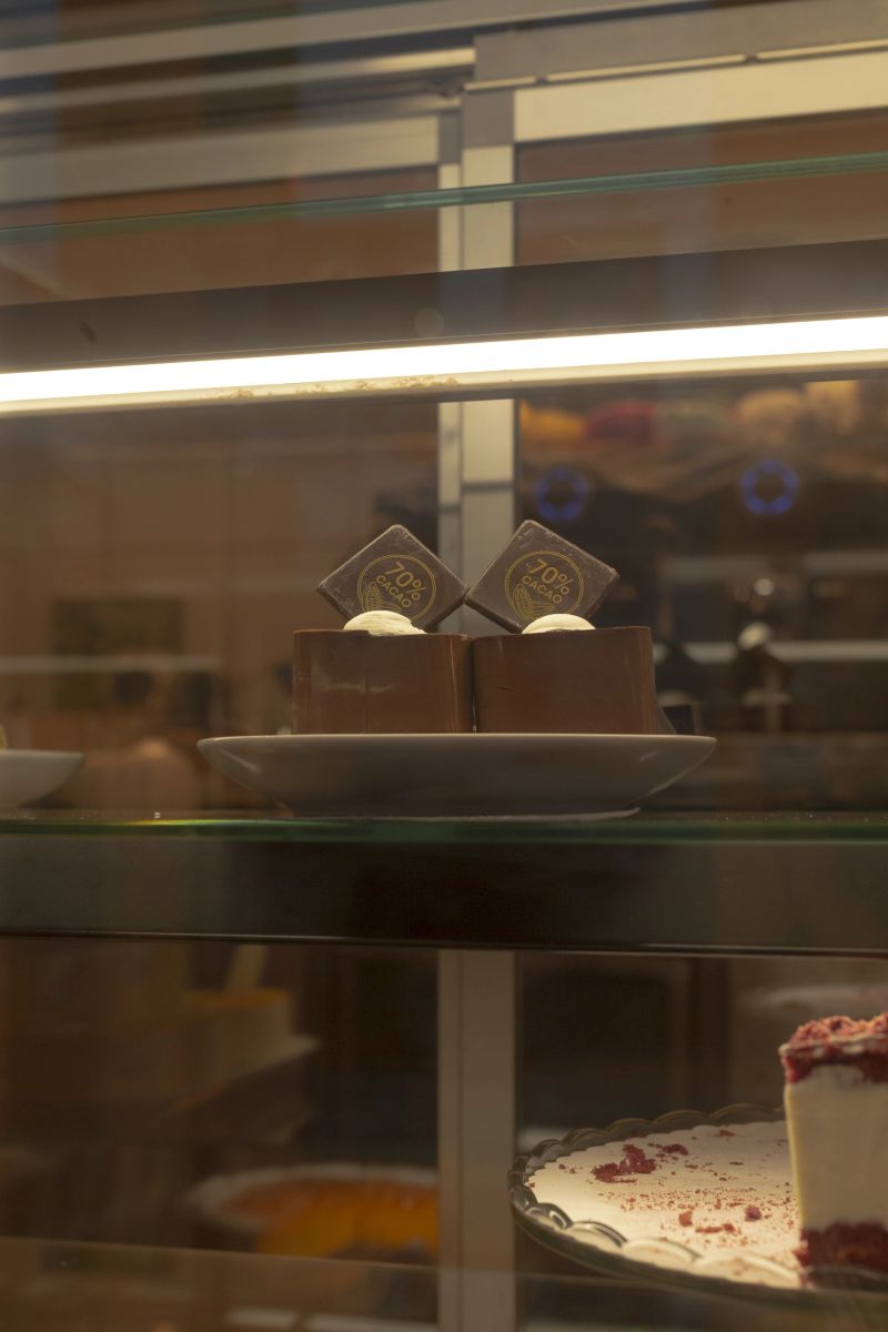 Pastry display inside restaurant interior, warm golden lighting illuminating glass case with sliced cake and strawberry garnish, reflective surfaces and labeled trays, cozy bistro vibe,