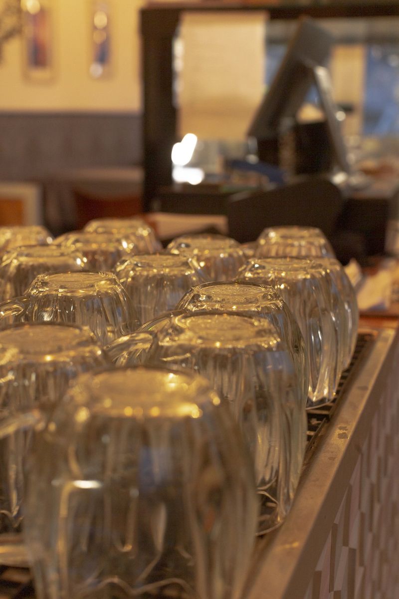 Glasses reflecting warm bar lights restaurant interior, closeup of glass rows with shimmer and bokeh, moody lounge atmosphere and polished counter reflections creating intimate setting