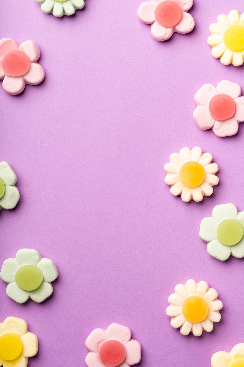 Assorted pastel flower-shaped gummy candies arranged on a purple background, creating a playful and sweet composition with empty space in the center