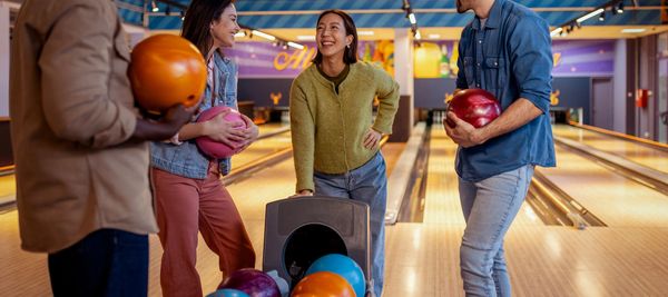 Four friends smiling and holding bowling balls at a bowling alley.