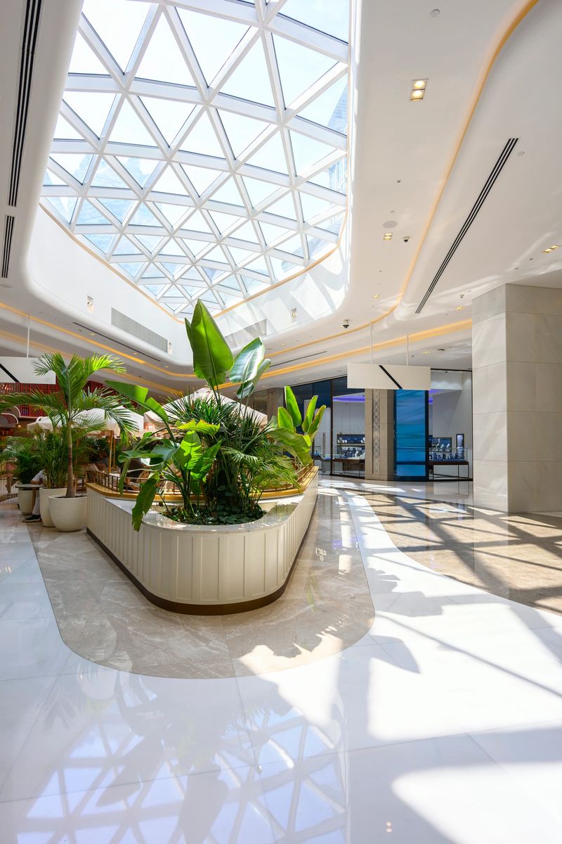 Large contemporary interior space showcasing a geometric glass skylight, white polished floors reflecting sunlight, and a central planter filled with lush tropical foliage, creating an urban oasis