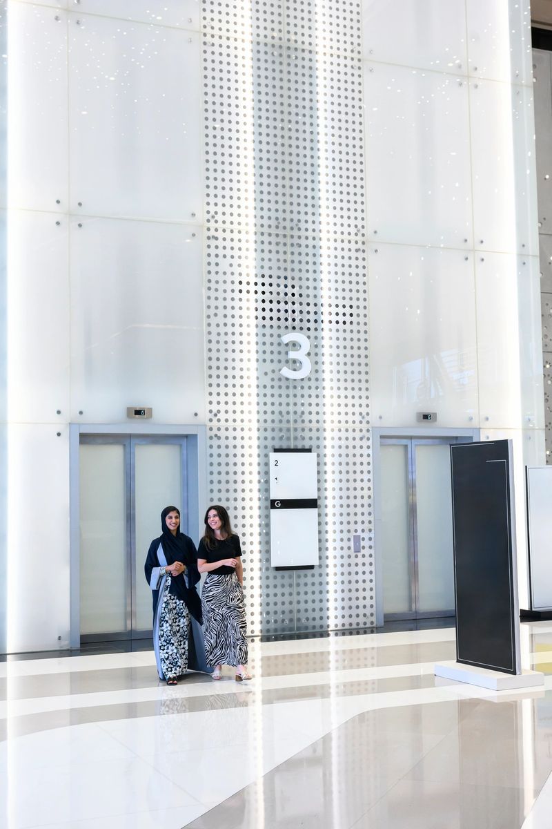Two businesswomen stand and converse in a bright, modern office lobby with sleek elevators, reflective floors and perforated white walls, projecting a clean, professional vibe
