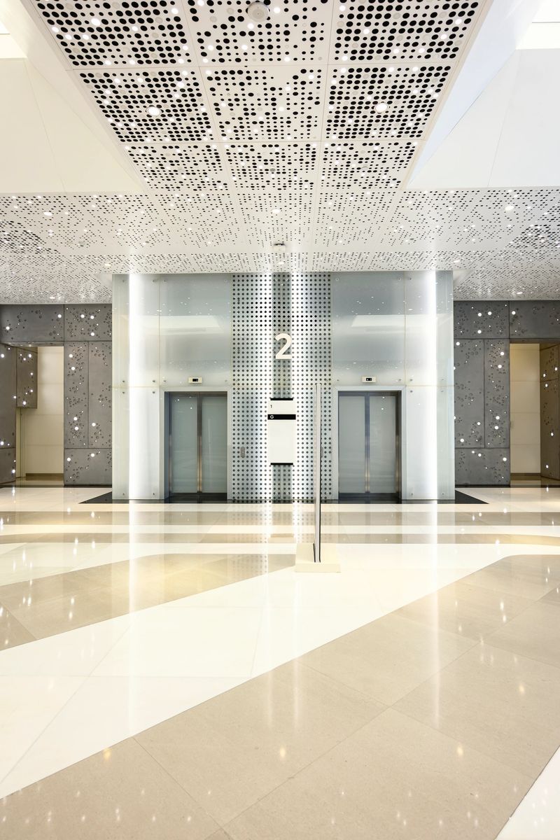 Contemporary lobby features two elevators, sleek glass panels, perforated ceiling, and polished floors reflecting natural light, creating a bright, spacious, and futuristic design
