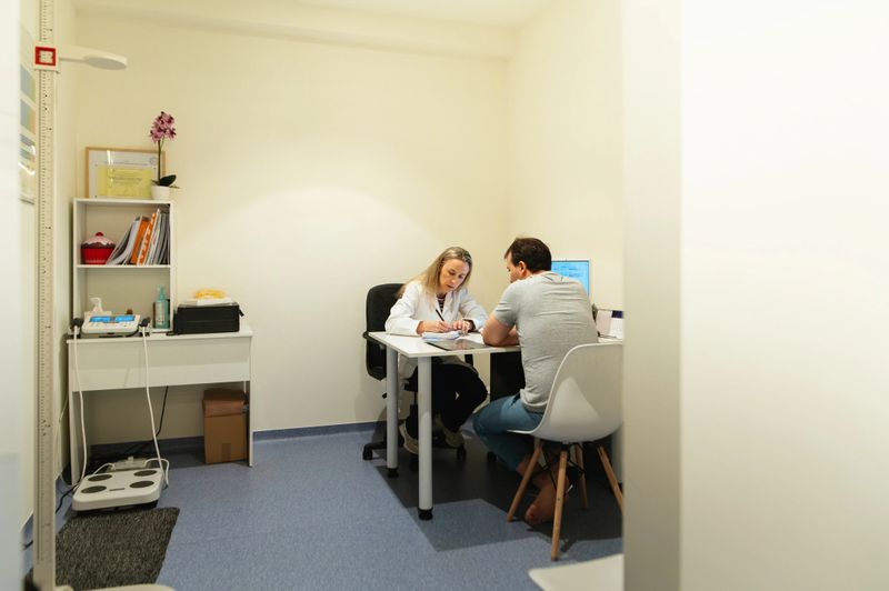 Female physician taking handwritten notes while speaking with an adult male patient seated across the desk in a private medical office.