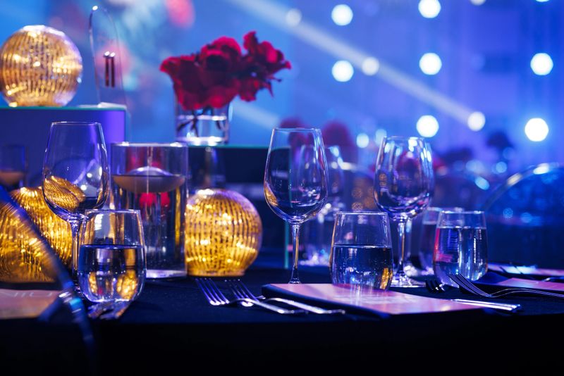 A banquet table features crystal glassware, red roses in a vase, and golden orb lamps. The background is filled with blue stage lighting and bokeh effects from a large venue.