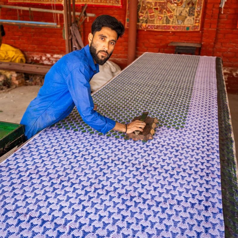 A bearded man wearing a bright blue shirt leans over a long table, using a carved wooden block to print repeating floral and leaf motifs onto a large piece of fabric. The cloth displays dense blue and green patterns. The worker appears focused as he presses the stamp, surrounded by a workshop setting with a red brick wall and hanging textiles in the background.