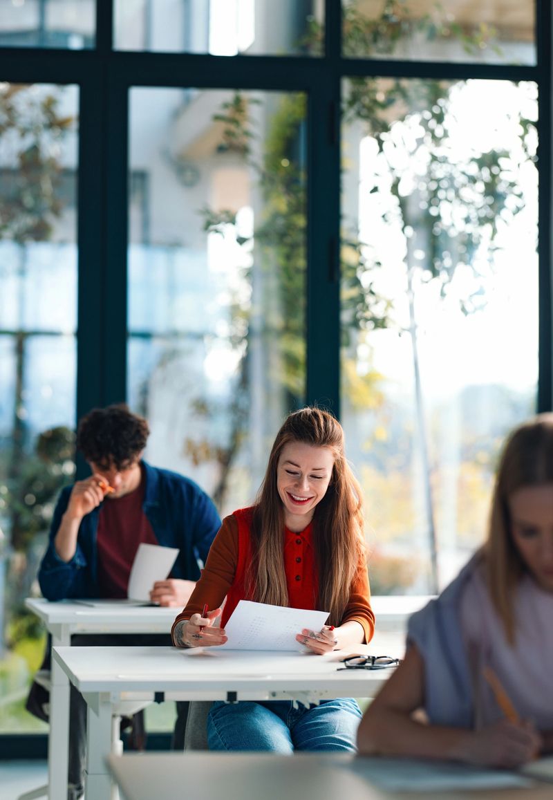 Smiling students sit together in a bright university classroom or high school study space, reading papers and taking notes during an exam or group study session that conveys focus and collaboration.