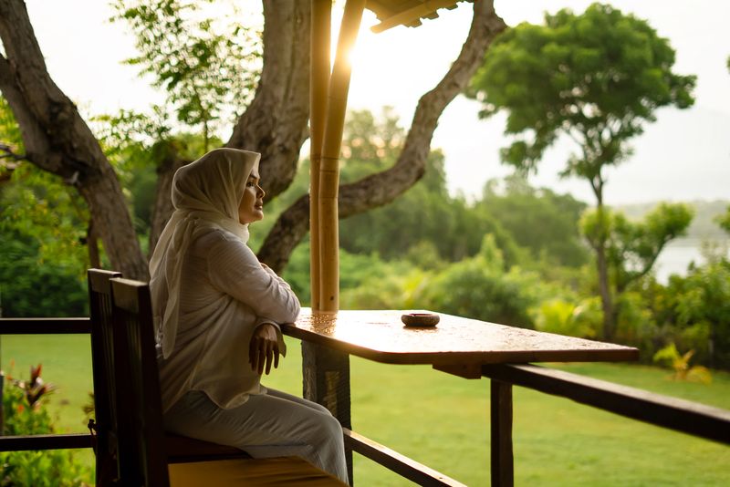 Young Muslim woman wearing a hijab sitting on a wooden terrace overlooking a lush green garden. She appears calm and reflective while enjoying a quiet outdoor moment in natural light. The scene represents peaceful lifestyle, mindfulness, relaxation, rural living, and spiritual serenity in a tropical environment.