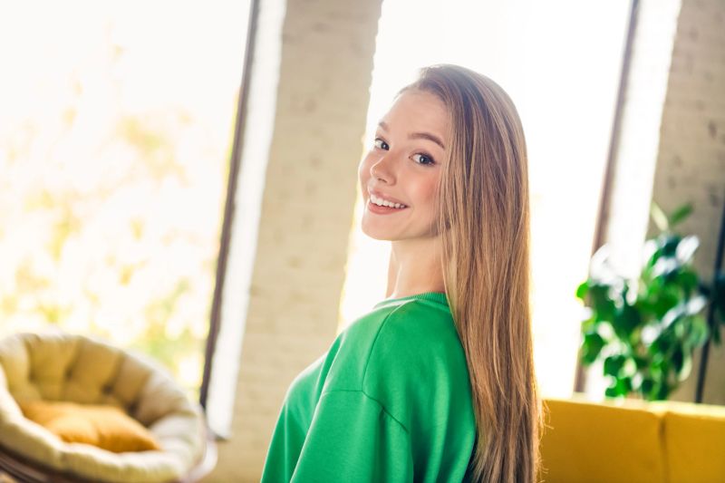 A happy young woman wearing a green sweatshirt smiles in a loft-style apartment, capturing the essence of comfort and relaxation.