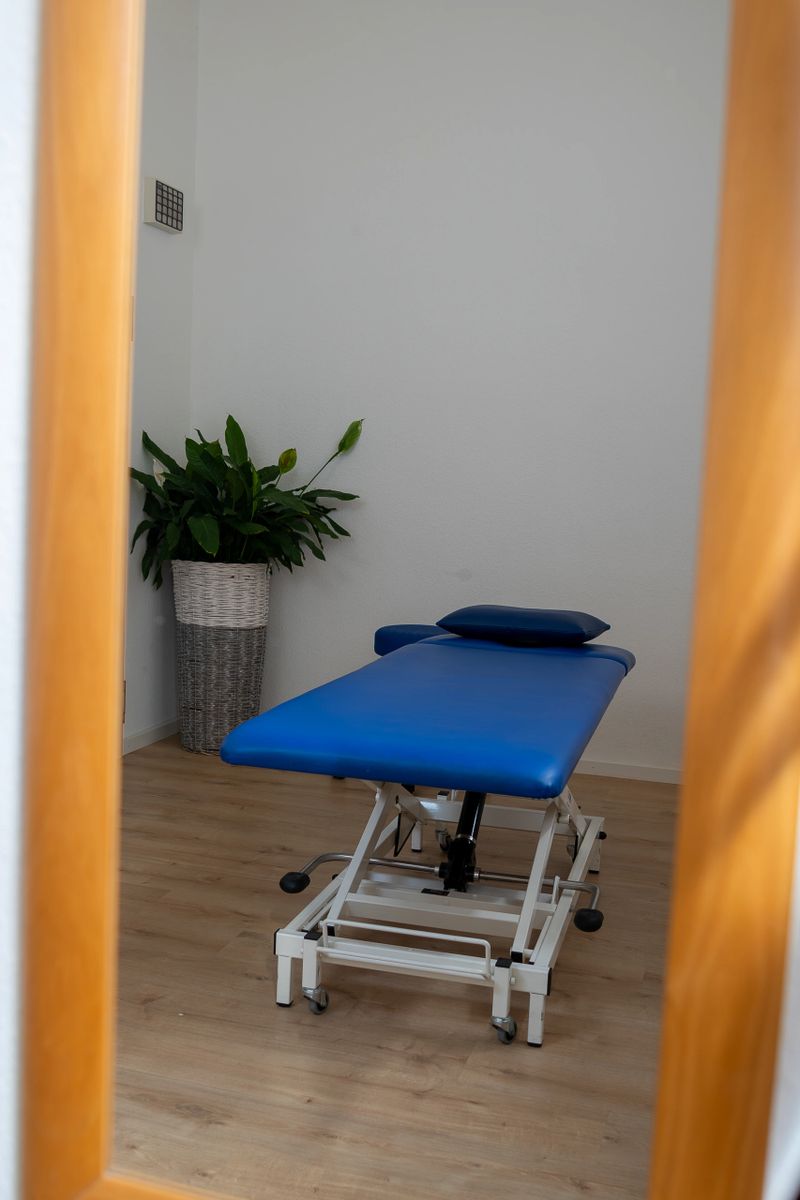 Empty blue treatment table in a physiotherapy practice therapy room with adjustable examination bed, potted plant and wooden floor, conveying a clean, professional and calming rehabilitation environment.