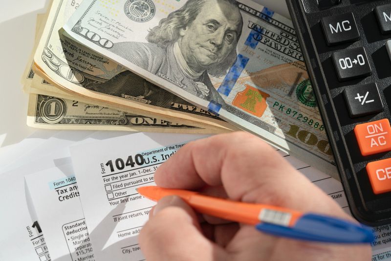 A close-up of a hand carefully filling out a US tax form 1040, surrounded by a stack of dollar bills and a calculator, under bright, clear lighting.