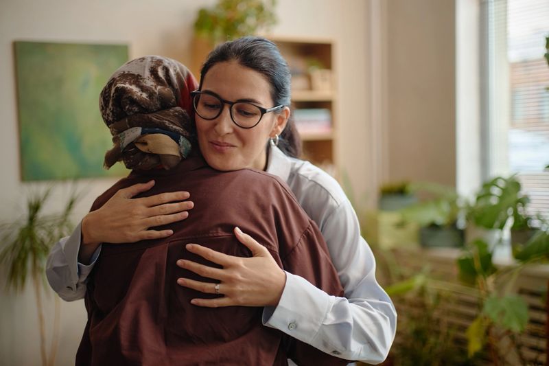 Middle aged Caucasian woman with glasses embracing middle aged woman in headscarf, showing support and empathy in counseling session, indoor setting with plants and bookshelf