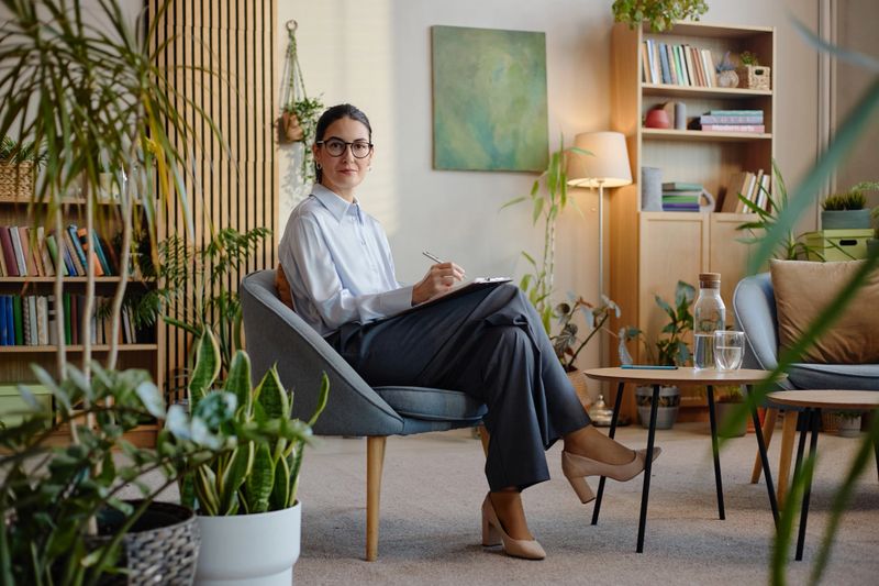 Portrait of young adult Caucasian woman sitting in modern office holding clipboard and pen, smiling and looking at camera, working as psychologist during therapy session