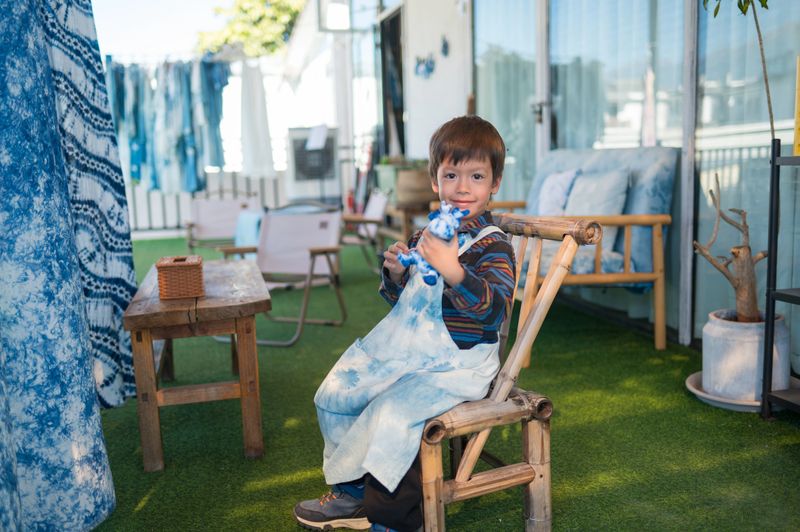 A joyful young boy proudly displays his handmade stuffed animal painted with vibrant tie-dye patterns. Colorful fabrics and a tie-dye apron surround him in a bright workshop setting, capturing the happiness and pride that come from creative textile crafting activities.
