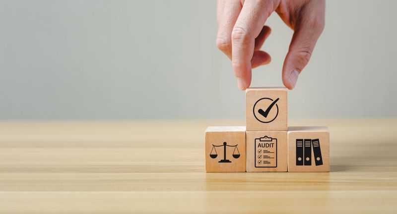 Hand placing wooden block with checkmark icon on compliance audit cubes featuring law balance, document checklist and binder symbol on desk, regulatory risk management concept