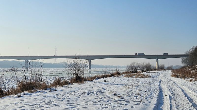 Side view of a long concrete bridge with several semi-trailer trucks crossing over a river filled with ice floes. A snow-covered riverbank with tire tracks is visible in the foreground under a clear sky. Concept of supply chain, winter logistics, and infrastructure maintenance.