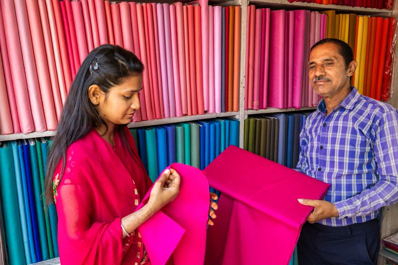Inside a brightly colored textile shop, shelves are stacked with neatly rolled bolts of fabric in a rainbow of hues. A man in a blue checked shirt holds a large magenta bolt while a young woman wearing a pink-red dupatta carefully examines and feels the cloth. The scene highlights vivid saturated colors, the texture of the material, and a moment of choosing fabric in a small retail setting.