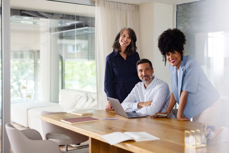 A diverse small business team collaborates around a table, smiling and reviewing work on a laptop.