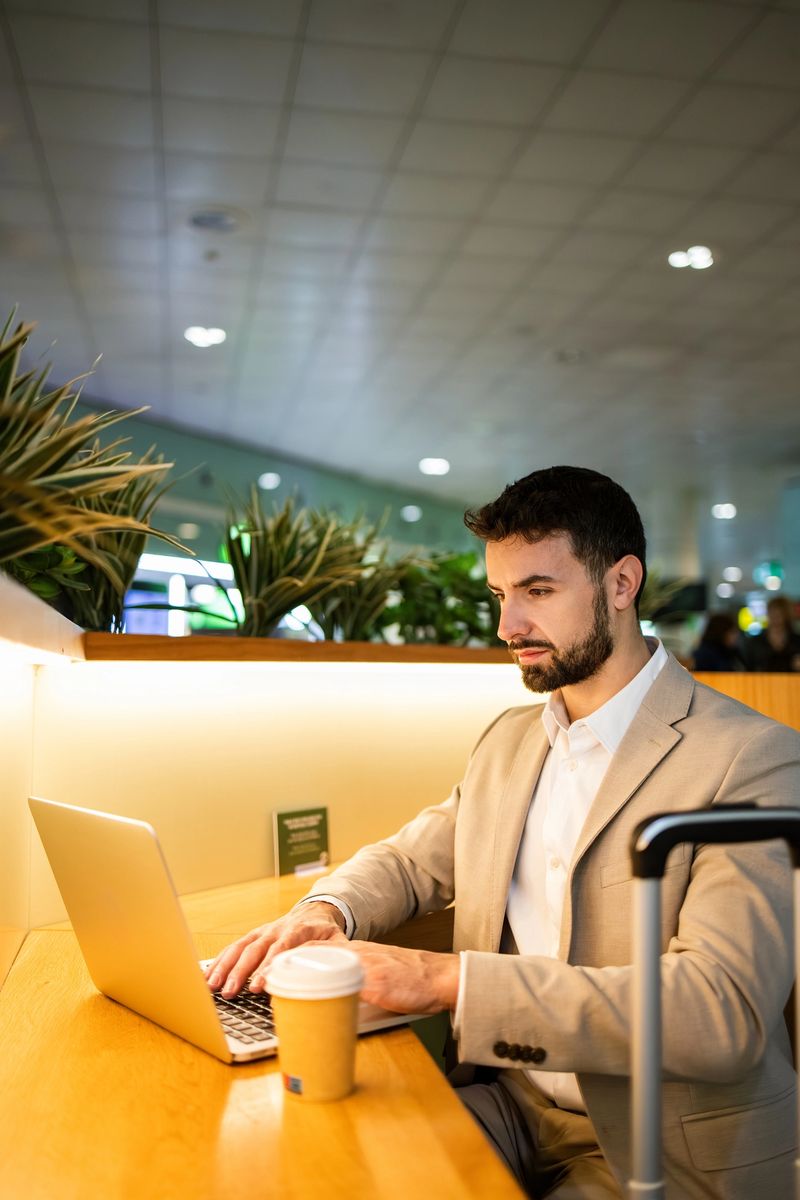 Businessman working on laptop in an airport lounge, typing on the keyboard with a coffee cup and luggage nearby