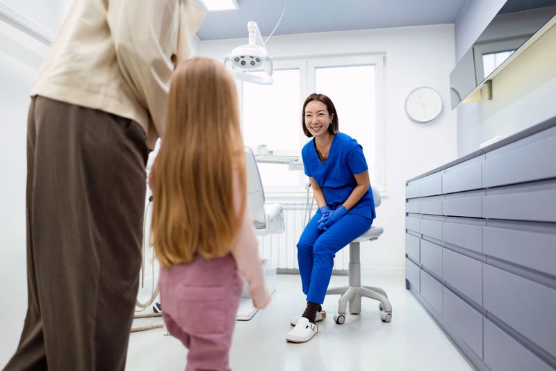 A friendly female dentist in blue scrubs greets a young girl and her parent in a bright dental clinic.