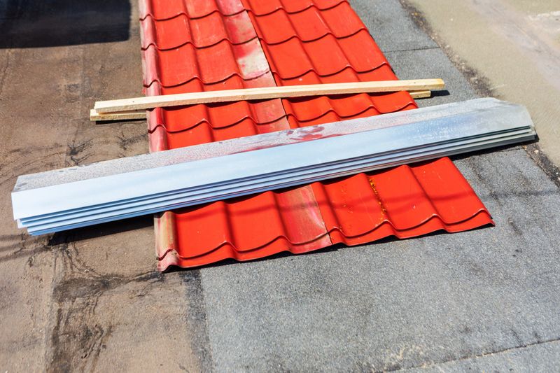 Section of a roof under construction shows bright red metal tiles laid out beside wooden planks and a sheet of galvanized steel. The materials are arranged on a flat surface, ready for installation