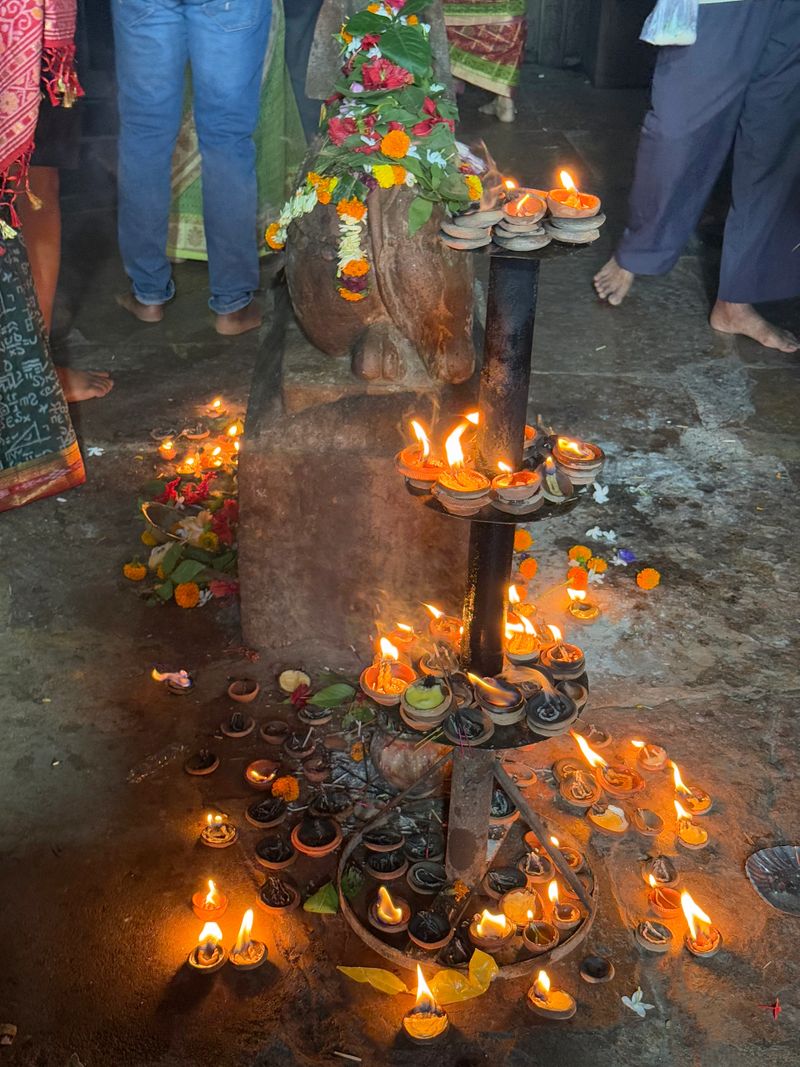 Lit oil lamps (diyas) surround a sacred stone idol decorated with flowers inside a Hindu temple, as devotees perform rituals and prayers. The scene captures spiritual devotion, traditional worship practices, and the warm glow of ceremonial lighting. Ideal for themes of religion, faith, rituals, Indian culture, and spirituality.