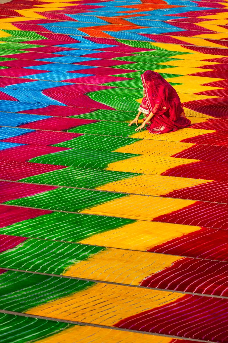 A person draped in a red sari crouches amid vast rows of brightly dyed fabrics spread out to dry. Repeating chevron-like bands of yellow, green, blue and magenta create a bold, rhythmic pattern across the scene, with stitched lines adding texture. The lone figure provides scale and a quiet, focused human presence against the intense, geometric colors.