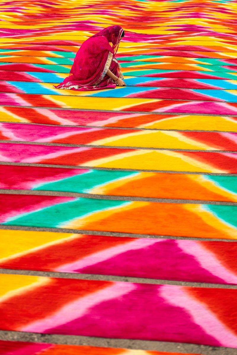 Rows of brightly colored, patterned rugs or fabrics are spread across the ground in repeating bands of pink, orange, yellow and turquoise. A woman in a magenta sari crouches among the strips, tending or arranging the textiles, adding a human scale to the vivid, sunlit composition. The strong perspective and rhythmic patterns create a striking visual of color, texture and craft.