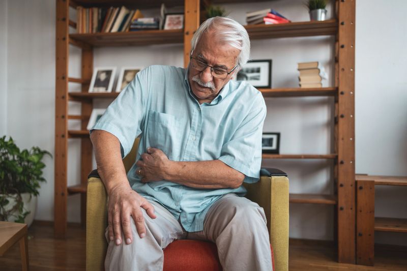 Senior man sitting in a chair at home, suffering from severe stomach ache or kidney pain. Health and medical concept