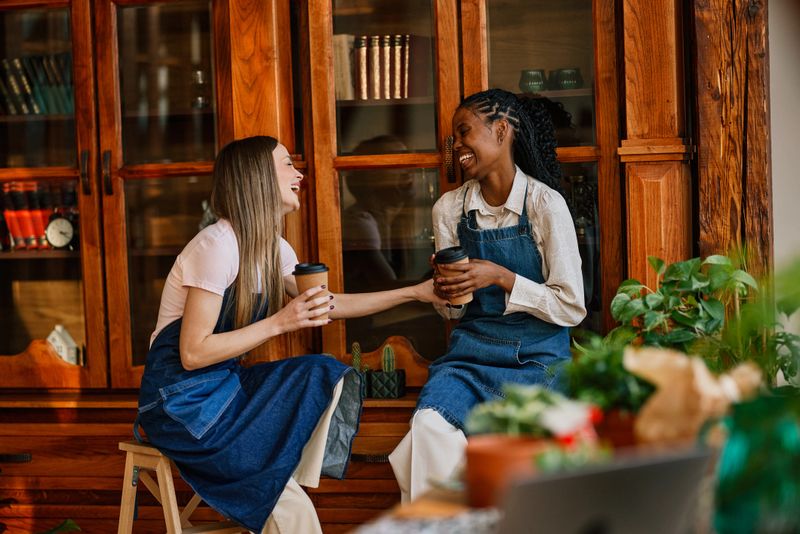 Two young women small business owners sit in a warm, wood-paneled shop talking and laughing over takeaway coffee among potted plants, showing friendship, connection and relaxed teamwork.