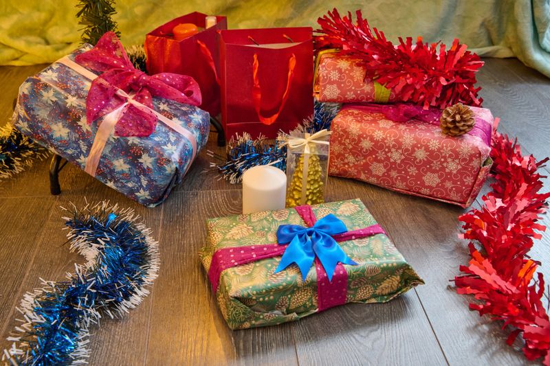 Wrapped presents in vibrant paper with ribbons, red gift bags, white candle, red and blue tinsel on dark wooden floor, cozy indoor environment, warm lighting